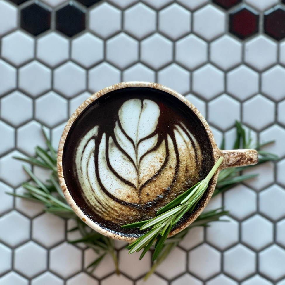 A cinnamon toast flavored hot latte in a porcelain cup on a black and white hexagon tile counter. The latte is finished in a rosetta shape and topped with sprig of rosemary.