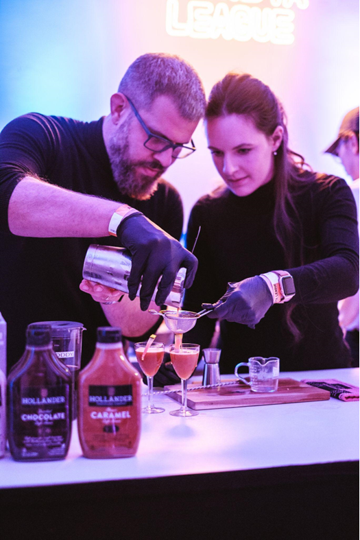 One male and one female barista, both wearing black turtlenecks and gloves, are creating craft beverages. One holds a strainer over a cocktail glass while the other strains liquid into it from a cocktail shaker.