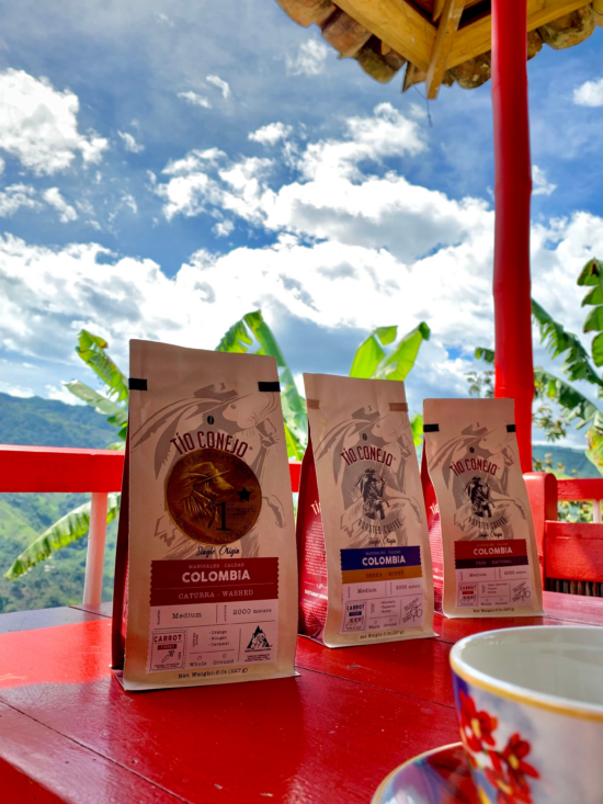 Wizerunek trzech torebek z kawą Tio Conejo siedzących na czymś, co może być czerwoną werandą.  Trzy różne kolumbijskie odmiany w papierowych torebkach kraft z różnymi kolorowymi etykietami.  Za nimi błękitne niebo z puszystymi chmurami i liśćmi palmowymi.