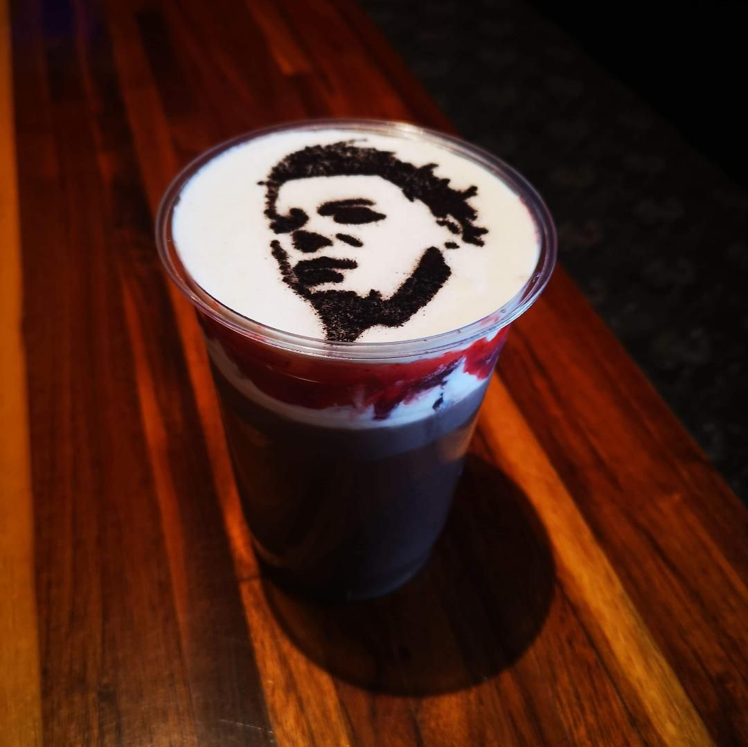 An iced coffee drink with foam on top of red "blood" sauce sits on a wooden tabletop. The profile of Michael Myers of the Halloween movie franchise is dusted on top in charcoal.