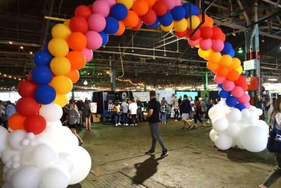 Um arco-íris de seis metros de comprimento feito de balões coloridos, com balões brancos formando um aglomerado de nuvens na parte inferior do arco.