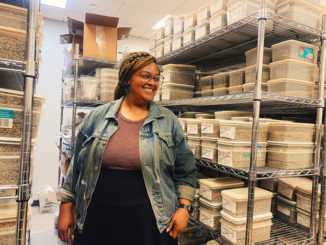 NKG Pace Partner Porttia Portis stands, smiling, in front of shelves full of boxes. Porttia has braids and glasses, wears black pants and a denim jacket over a brown top.