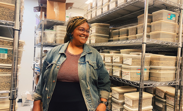 NKG Pace Partner Porttia Portis stands, smiling, in front of shelves full of boxes. Porttia has braids and glasses, wears black pants and a denim jacket over a brown top.