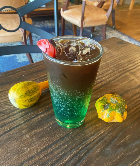 A glass filled with green melon tonic water on bottom, espresso and ice floating on top, with a little gummy fish sticking out of the drink. Gourds are lying on the wood table beside it.