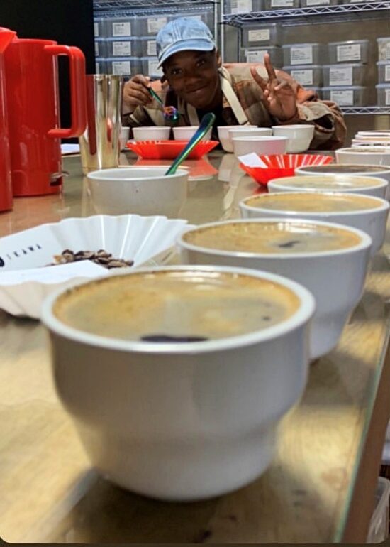 Jay flashes a peace sign while bending over a cup during a cupping session. They are wearing a denim baseball cap. In the foreground is a close-up of ceramic cupping cups.