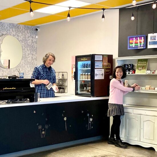 Tran and a coworker at Lava Java. Tran stands at a white cupboard, organizing some retail items. She is turned towards the photographer, laughing. She wears a frilly skirt with tights and boots, and a sweater. Her coworker behind the bar is pouring milk from a metal pitcher into a paper cup. To his left is a Strada machine.