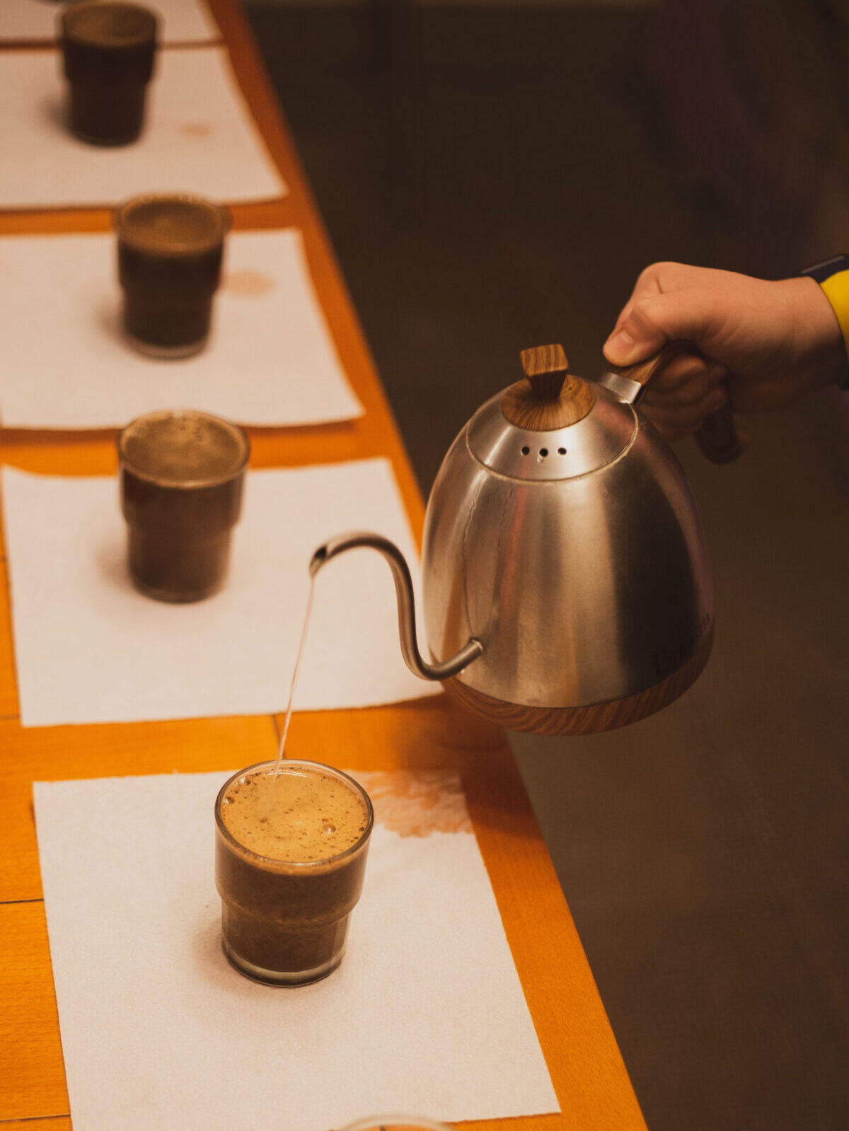 En hånd hælder varmt vand fra en svanehalskedel over kaffegrums i klare glaskopper.