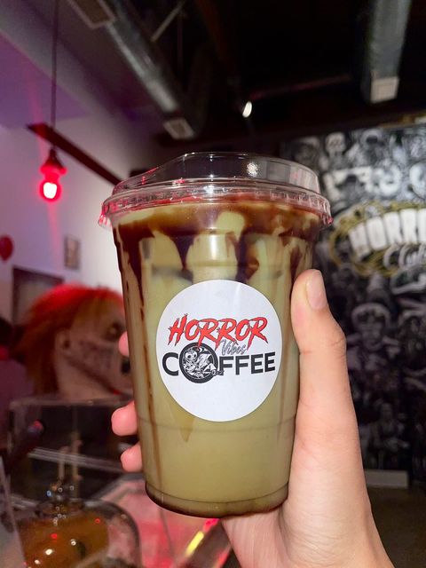 A hand holds up an iced mocha drink in front of the black and white horror themed wall at Horror Vibes Coffee. There is a red light bulb and scary zombie mask in the background.