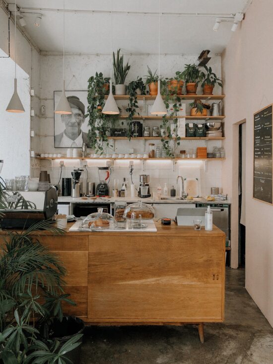 The Scandinavian minimalist inspired counter at Hello Kristof features a wooden bar on mid-century shaped peg legs, glass cloches covering pastry offerings, open shelving on the back wall, a large black and white photo of an elderly man, an espresso machine, trailing plant leaves, and a black chalkboard with a written menu.