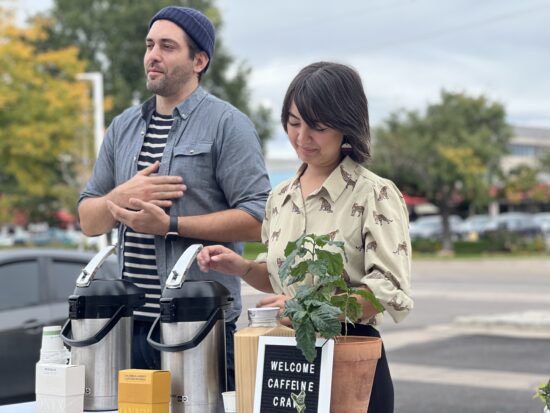 John wears a beanie and chambray shirt over stripes. Kristi wears a button-up shirt with cheetahs printed on it and black jeans. In front of them on a table is a sign that says welcome caffeine crawl. Also are coffee boxes, urns, and a potted plant.