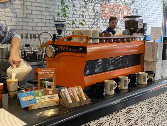 A bright orange espresso machine sits on the work counter at Ozo. Ozo branded mugs are lined up in front of it. A barista pours shots behind the bar.