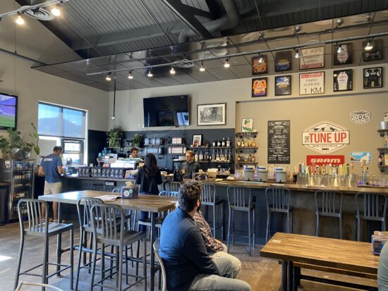 The bar inside Tune-Up Tavern. There are liquor bottles and bicycle signage along the wall. There are tall barstools in front of the counter and tall top tables throughout the room.