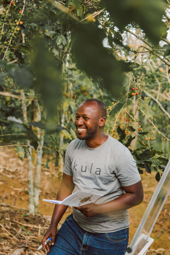 A Rwandan man holding a booklet smiles at someone off screen. He wears a grey Kula tee and blue jeans.
