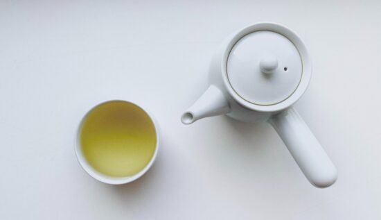 A plain white porcelain teacup with no handle is full of brewed white tea, a pale yellow-green color liquid. Beside the cup is a small white teapot with a lid and a handle on the side, similar to a pot handle. 