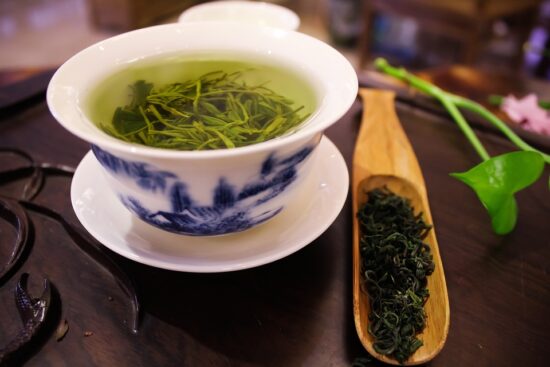 In a white and blue china teacup on a white saucer, there is green loose leaf tea brewing. Beside the cup is a wooden scoop with more dried green tea leaves. Both are on top of a carved wooden board.