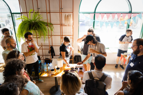 Two participants are cupping the  Natural Colombia from Finca La Rivera. The tables are covered in ceramic cupping vessels, water bottles and paper towels. Spectators stand around the café watching. 