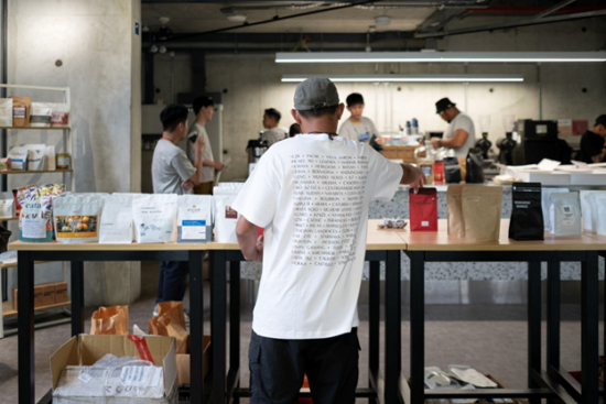 Back view of a man at the cupping table arranging many bags of display coffees brought by Singapore roasters for the event. He wears a white tee and grey baseball cap.