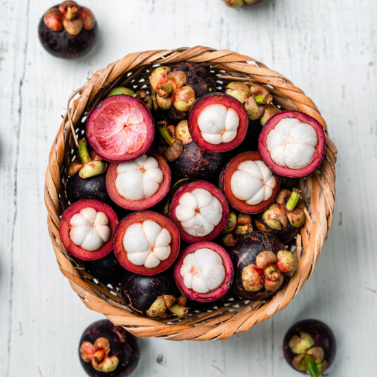 A hand-wpven round basket is filled mangosteen, some with peel intact, others with the white flesh inside visible. The skins are dark purple, with green leaves by the thick stems at the top. The flesh looks similar to the pips of an orange, only it is bright white.