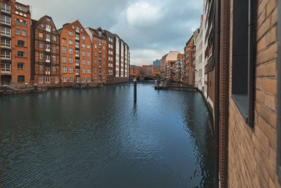 Brick buildings with pointed roofs tower over the water of the Nikolaifleet, a canal in Hamburg. The buildings go right up to the edge of the water.