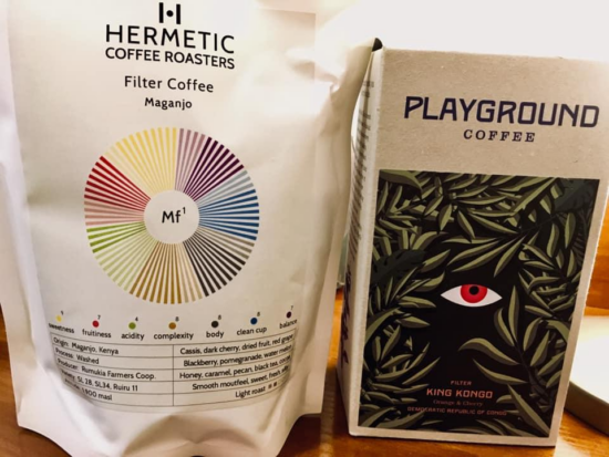 On the left is a white coffee bag from Hermetic Coffee Roasters labeled "Filter Coffee Maganjo." It has a multicolored flavor wheel rating the level of the various flavors in the coffee. On the right is a brown paper box of Playground Coffee beans labeled "Filter King Kongo." It features an image of a single red eye on a black background, with dark green leaves surrounding.