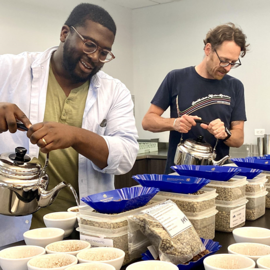 Charles e um colega despejam água de chaleiras de prata em vasos de cerâmica para experimentar o café.  Diante deles, sobre a mesa, estão muitos copos, caixas plásticas com grãos de café e bandejas.
