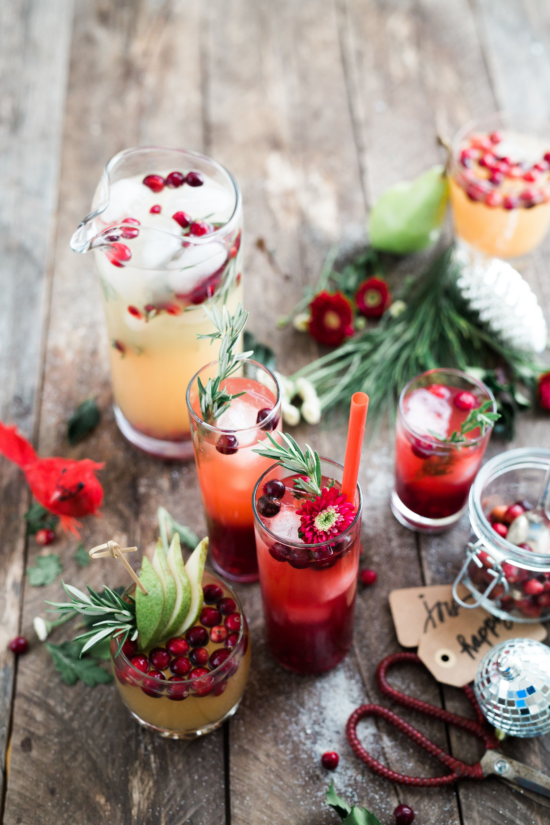A festive red and green seasonal display of drinks on a wood table with cranberries, flowers, greenery, apple slices on skewers, rosemary sprigs and pomegranate. Adorning the table is a decorative cardinal figurine, fake snow, flowers, and ribbon.