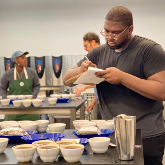 Charles takes notes in a notepad about the coffee he is tasting. Many ceramic cupping vessels lie before him on a metal table, complete with coffee trays.