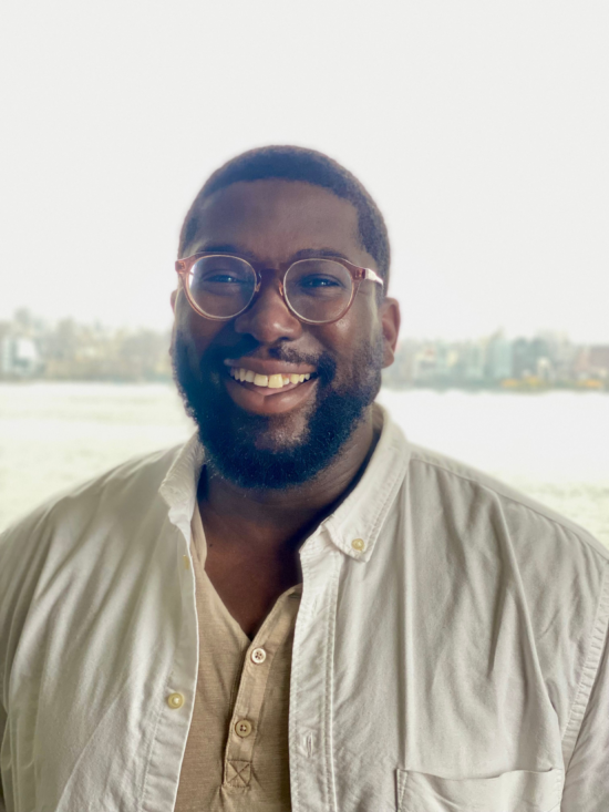 Charles smiles for the camera, wearing a white button-up shirt and glasses. In the background is a large body of water and a city skyline.