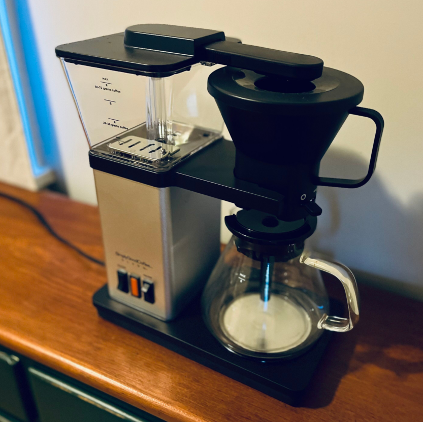 A top view of the Olson Coffee Brewer. It looks similar to automatic pour-over machines with a top cone and glass carafe at the bottom on one side. The other size stores water  and a heating element inside a rectangular metal tower, and has a bar to deliver water from the tower. 