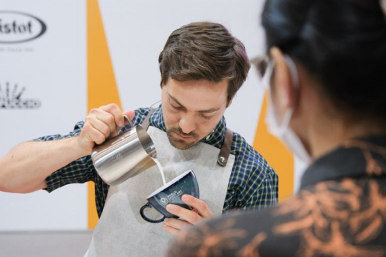 Johannes pours in a latte art competition. He has short hair and a close beard. He wears a linen apron with smooth leather neck loop. He is pouring milk from a silver pitcher into a black ceramic latte mug.