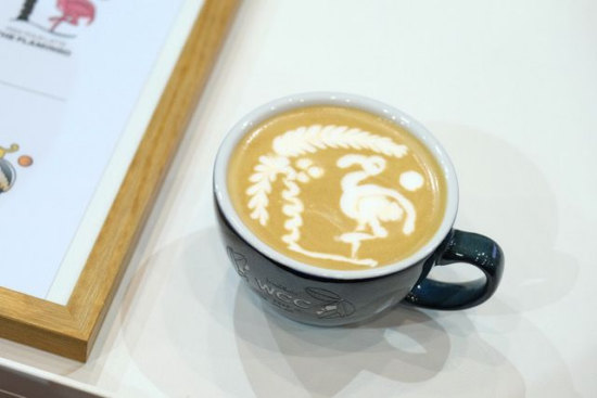 Latte art of a flamingo and a coconut palm tree. There is a little sun in the background of the milk and the flamingo is standing on one leg.