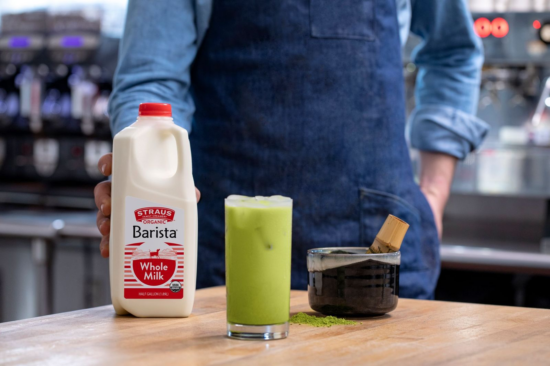 A barista stands in front of a bottle of milk with one hand in their pocket. There is a matcha bowl and whisk in front of them, and an iced matcha in a glass.