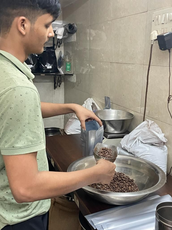 A man with a mustache scoops roasted coffee beans out of a large metal bowl into a plain silver coffee bag.