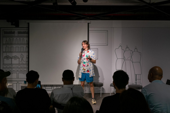 Matt Winton speaks before an assembled audience. He is holding a microphone in one hand, gesturing to the audience with the other. He is wearing bright colors with a whimsical dinosaur print tee under an open button-up shirt.