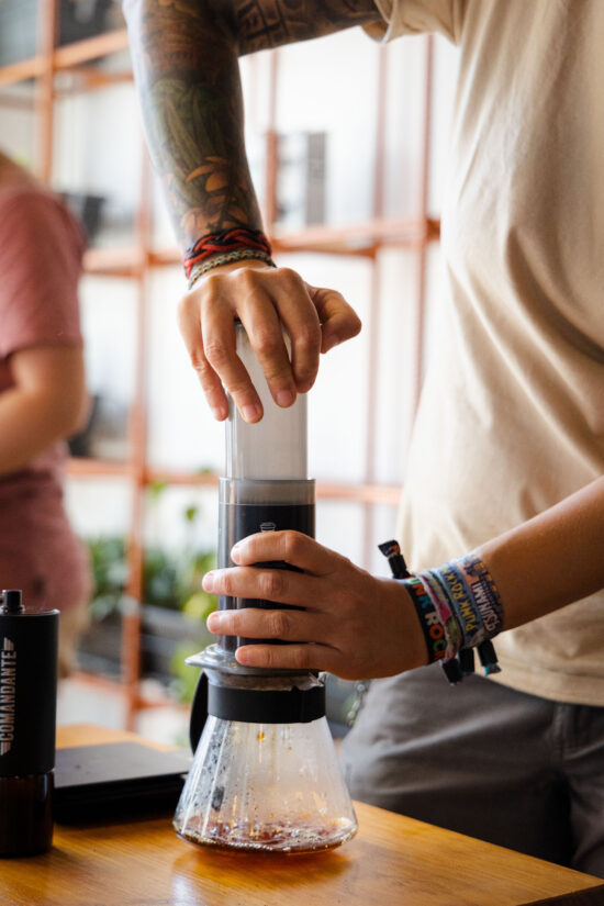 Someone pushes down the plunger of an AeroPress to brew coffee for the competition. They are wearing a white t shirt and stacked bracelets at the Portuguese Aeropress Championship.