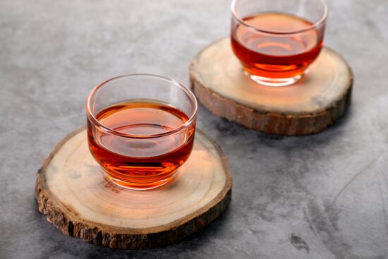 Two clear glass cups of black tea rest on coasters made of unfinished wood sliced out of the limb of a tree. Bark is still visible around the edges.