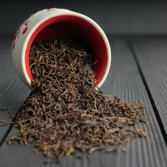Some fermented tea leaves spill from a decorative white jar with red interior onto the surface of a table.
