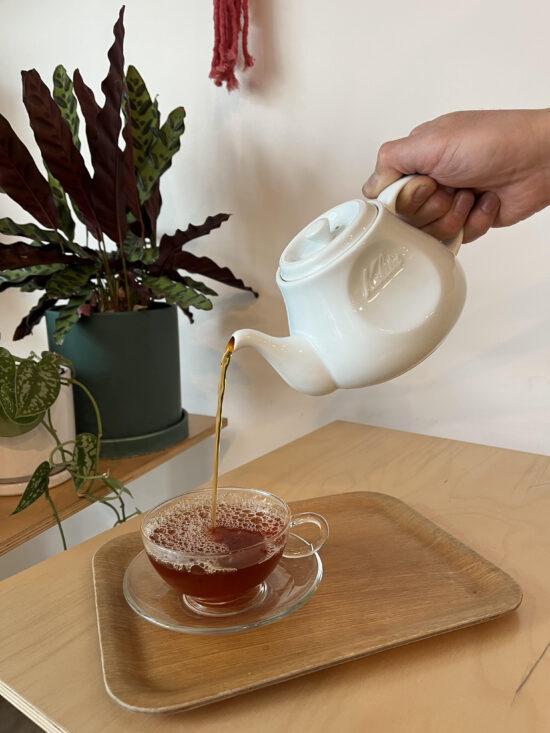 A person pours tea from a white teapot into a clear glass cup and saucer placed on a wooden tray.