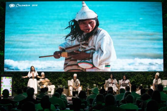 En kvinde henvender sig til publikum, mens en indfødt colombiansk kvinde i en keglehat på skærmen bagved holder en majskolbe.