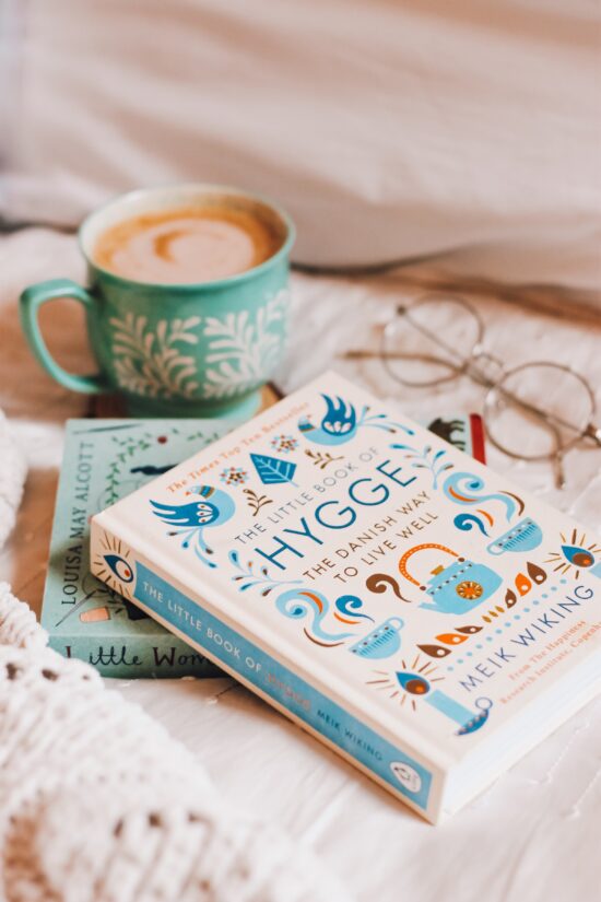 Two books arranged on a table with a blue and white mug and a pair of spectacles. One book is Little Women by Laura Ingalls Wilder; the one on top is Hygge: The Danish way to live well, by Meik Wiking. It is covered in traditional Nordic illustrations of birds, a teapot, a candle, greenery.
