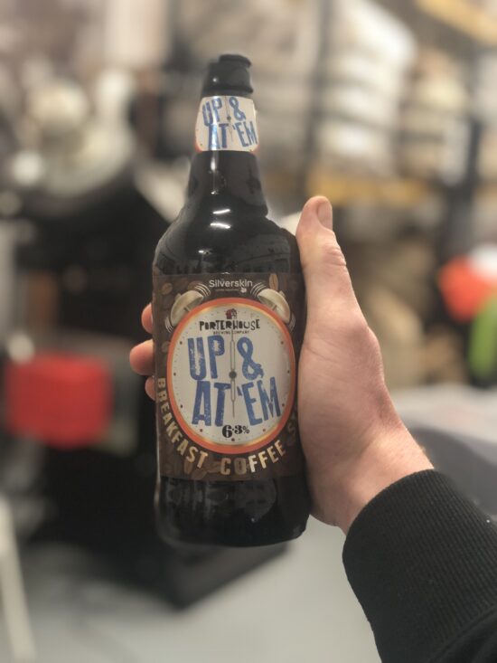 A hand holds up the Up and at em breakfast coffee stout. The bottle is brown glass, with an alarm clock complete with bells at the top making the label. The hands on the alarm clock read 6 o'clock. The Portehouse logo rests on the top of the clock and it is labeled 6.3% ABV.