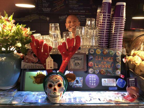 A man smiles behind the counter at a cafe. There is a sugar skull with reindeer antlers on the counter, stickers galore on the back of the espresso machine, and a bouquet of pink flowers beside it.