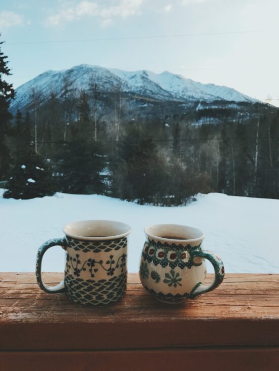 Two blue and white ceramic mugs of coffee sit on a wooden ledge overlooking a yard full of snow and evergreen trees several yards away. In the background, snow-capped mountain peaks.