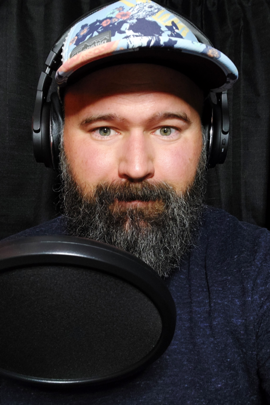Host of Roast West Coast, Ryan has a big beard and wears a floral patterned ball cap. He is seated in front of a large round microphone screen and wears headphones, smiling.