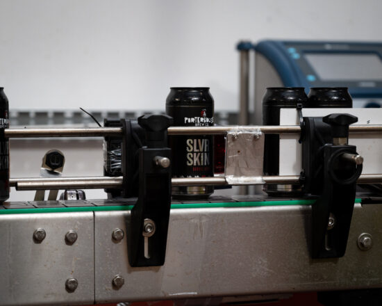 Showing part of the assembly line of the coffee stout beer cans. Cans are on a belt going past a short metal railing held in place with screws and clamps.