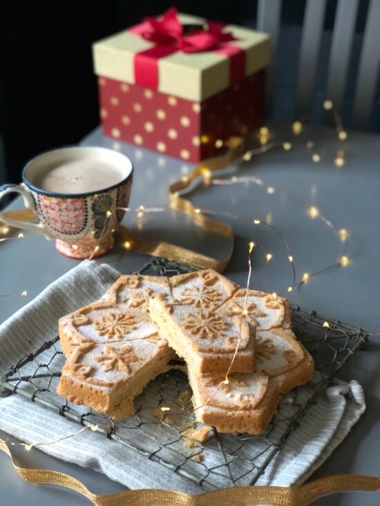 A Christmas present, a warm drink in a festive mug, and shortbread cookies shaped liked snowflakes on a white tablecloth with tiny fairy lights on string.