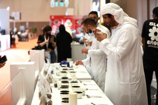 Three men in white, some with head coverings, sample coffees out of ceramic cupping vessels. They are standing at a long table under a white table cloth. Cupping vessels, coffee bags and spoons are neatly arranged down the length of the table.