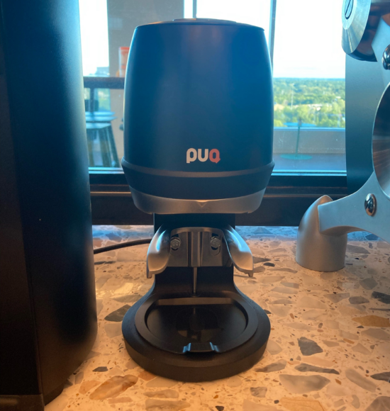 The Puqpress Q2 model, a small black contraption with silver rests for the portafilter and a dome on top that holds the pressing mechanism. The base is flat with a removable cover for catching grounds. It is labeled PUQ with the P and U letters in white and Q in orange.