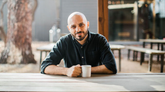 Chris is host of Keys to the Shop, has a shaved head, wears a button up shirt and hold a mug of coffee in one hand. He is seated at an outdoor patio table with a large tree in the background.