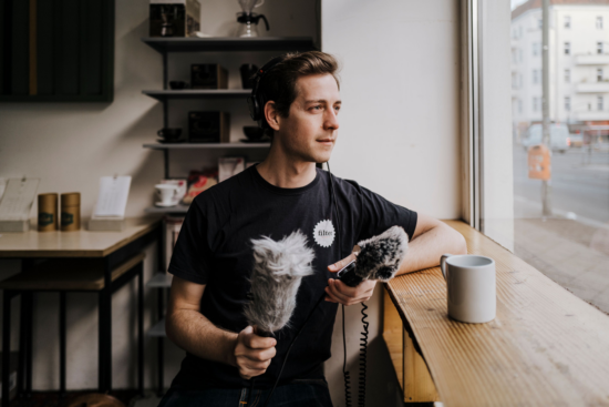 Filter Stories host James holds a fuzzy-covered microphone and wears headphones. He is seated at a counter looking outside a window at the city.